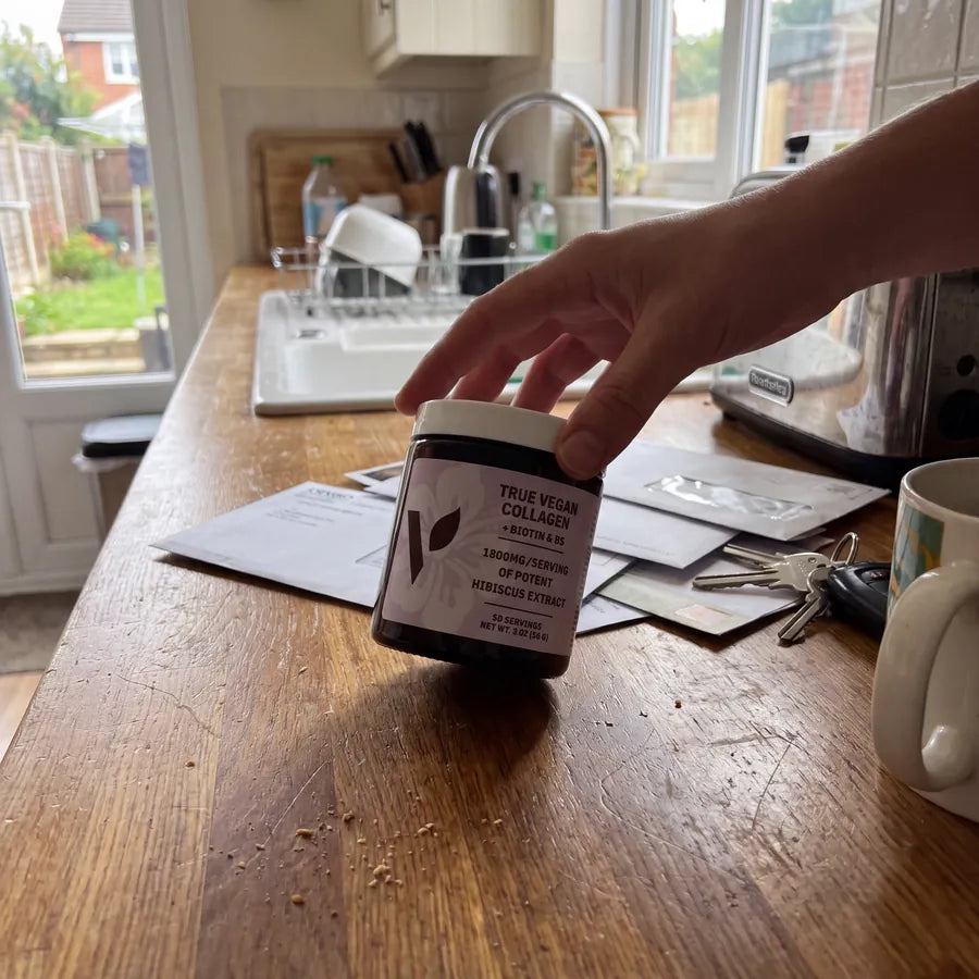 Hand picking up a jar of True Vegan Collagen from a kitchen counter with keys and letters