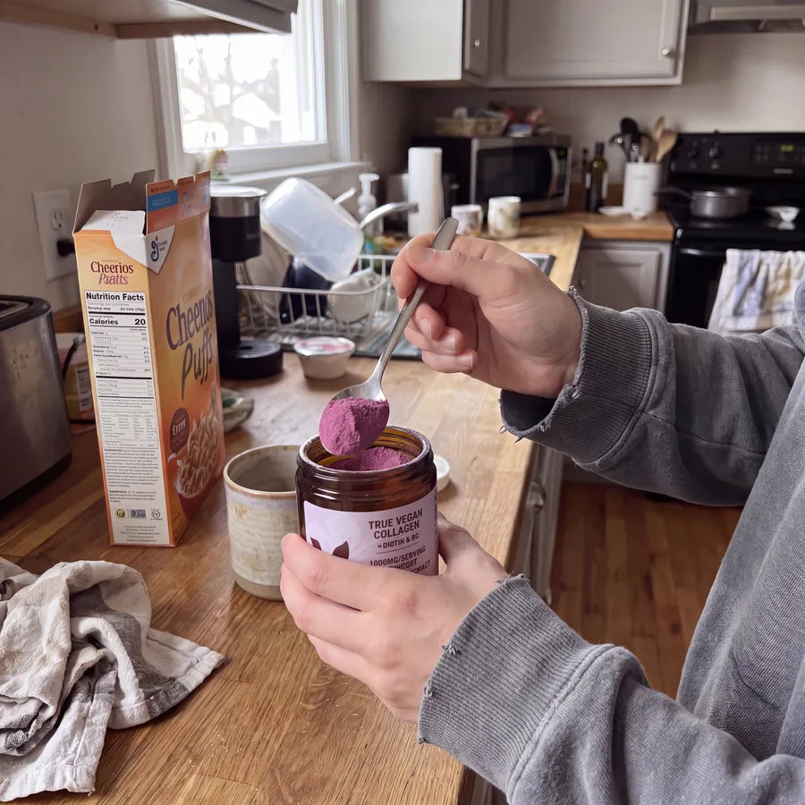 Hands spooning purple True Vegan Collagen powder from the jar at a kitchen counter