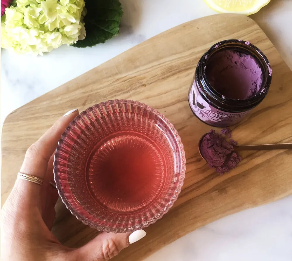 Hand holding a glass of pink hibiscus drink beside an open jar of True Vegan Collagen
